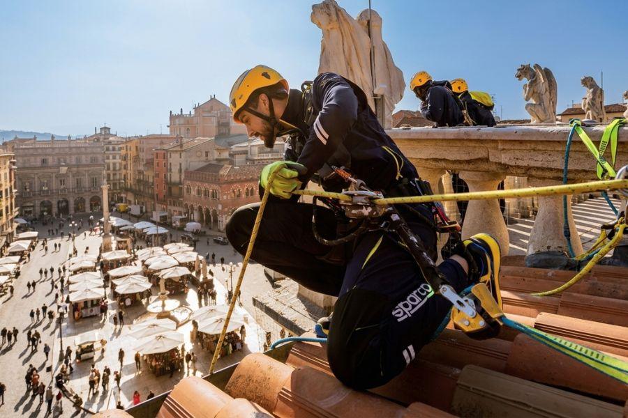 Un operaio specializzato, equipaggiato con DPI completi (casco giallo, imbracatura di sicurezza, corda di posizionamento con discensore, guanti protettivi e scarpe antinfortunistiche Sparco), è immortalato in una manovra tecnica di discesa in corda doppia. L'immagine, scattata da una prospettiva in quota, lo mostra mentre opera in sicurezza su una copertura in tegole di terracotta di un edificio storico. 